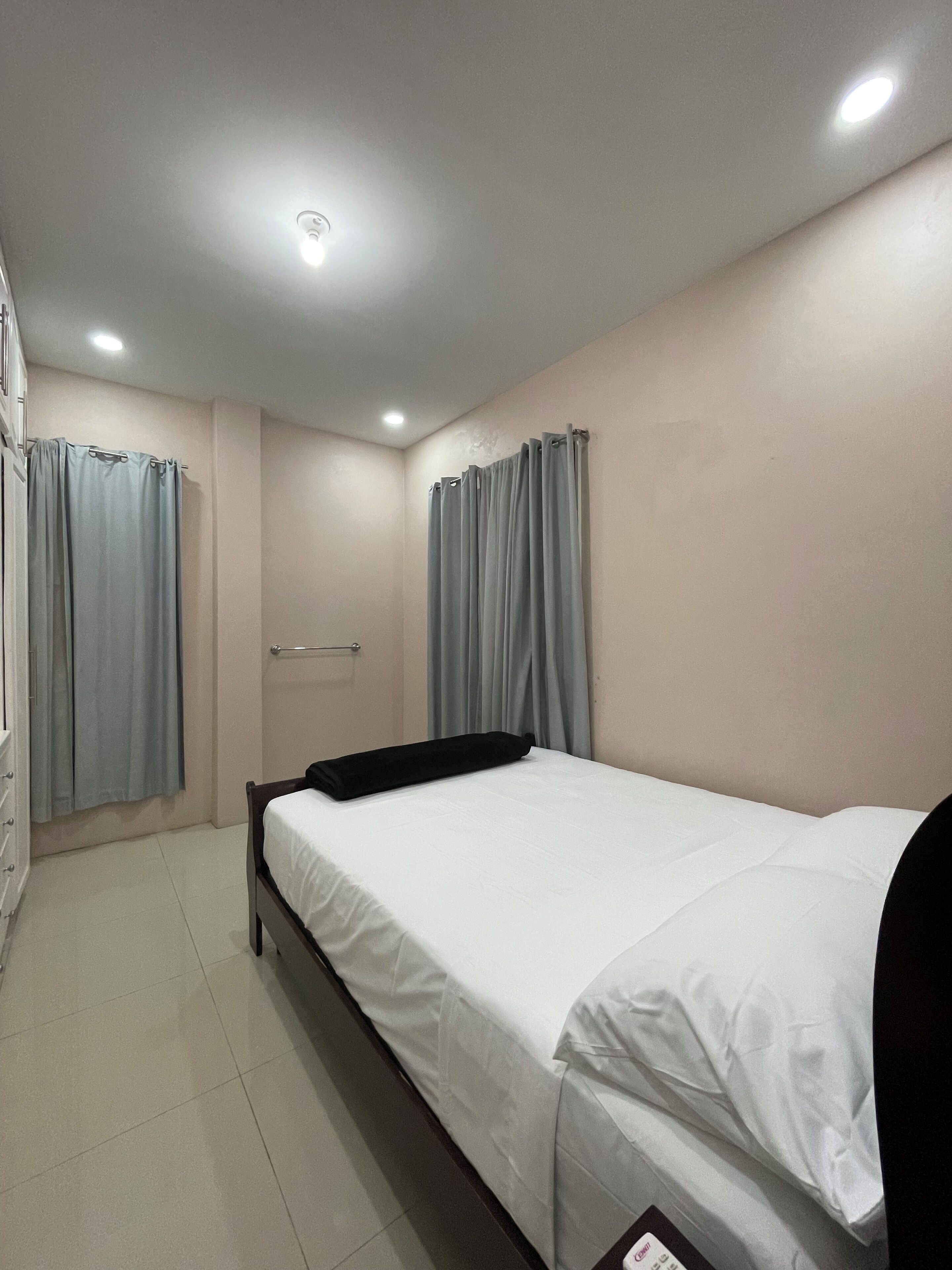 Un lit simple avec un matelas blanc et une tête de lit noire, dans une pièce aux murs beige, aux rideaux gris et au sol carrelé.