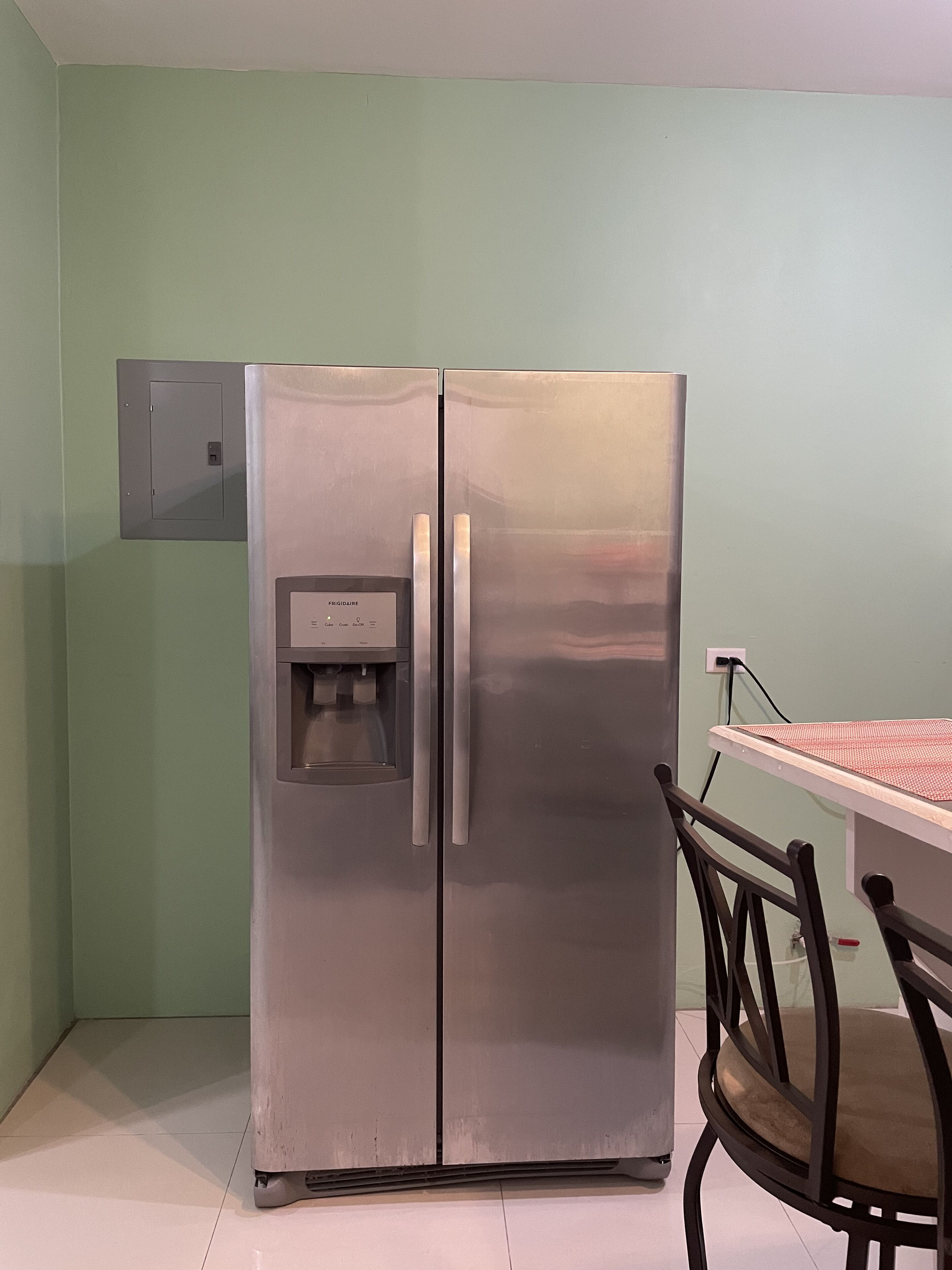 A stainless steel refrigerator with a water dispenser, a wooden chair, and a table with a red and white checkered tablecloth.