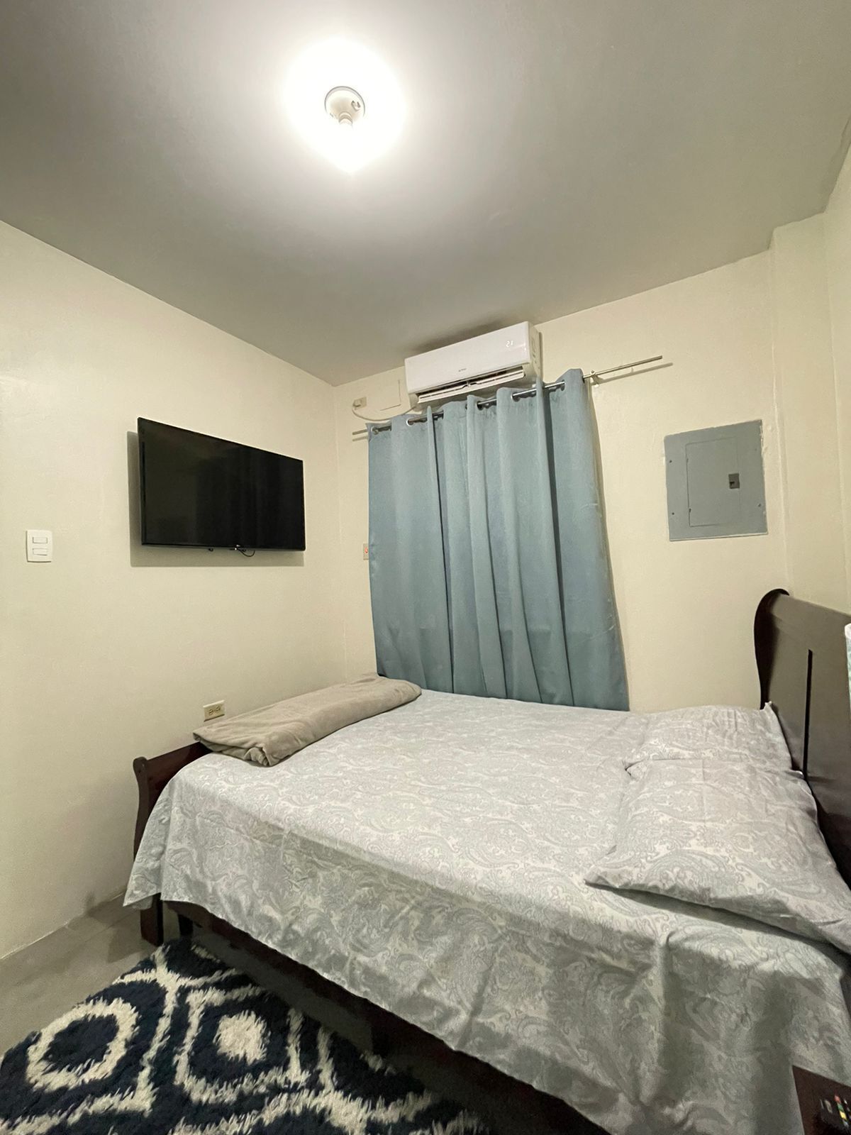 Un dormitorio con cama, televisor de pantalla plana montado en la pared, equipo de aire acondicionado y toma de corriente.
