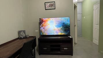 Ein Zimmer mit einem Holztisch, einem schwarzen Bürostuhl, einem Flachbildfernseher auf einem dunklen Holzständer und einem gerahmten Bild an der Wand.