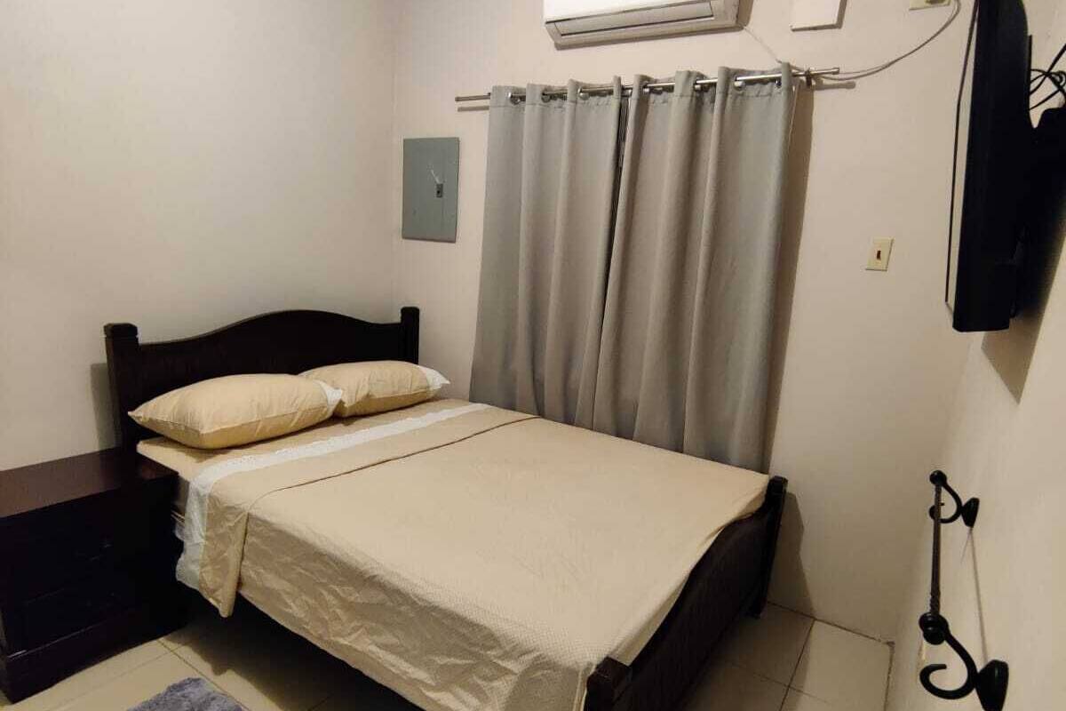A bedroom with a bed, a nightstand, an air conditioning unit, a wall-mounted TV, and a grey rug.