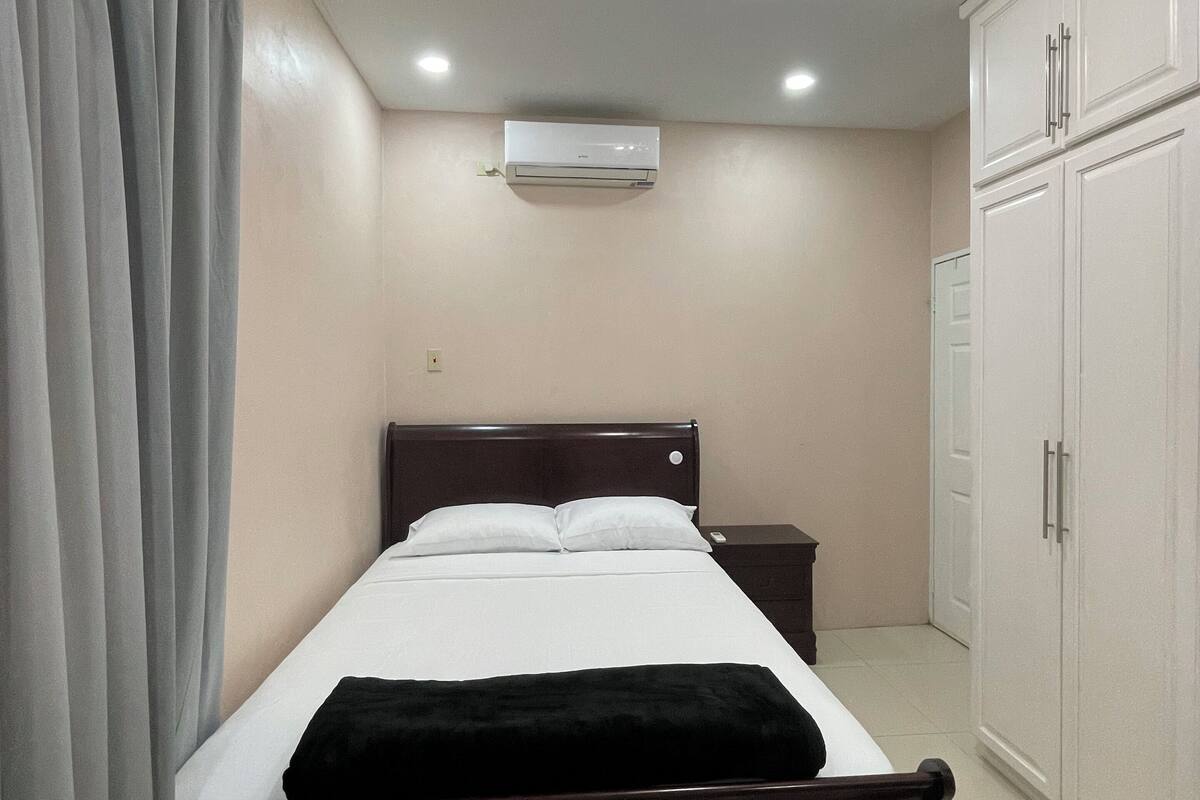 A hotel room with a single bed, a bedside table, a wardrobe, and an air conditioning unit.