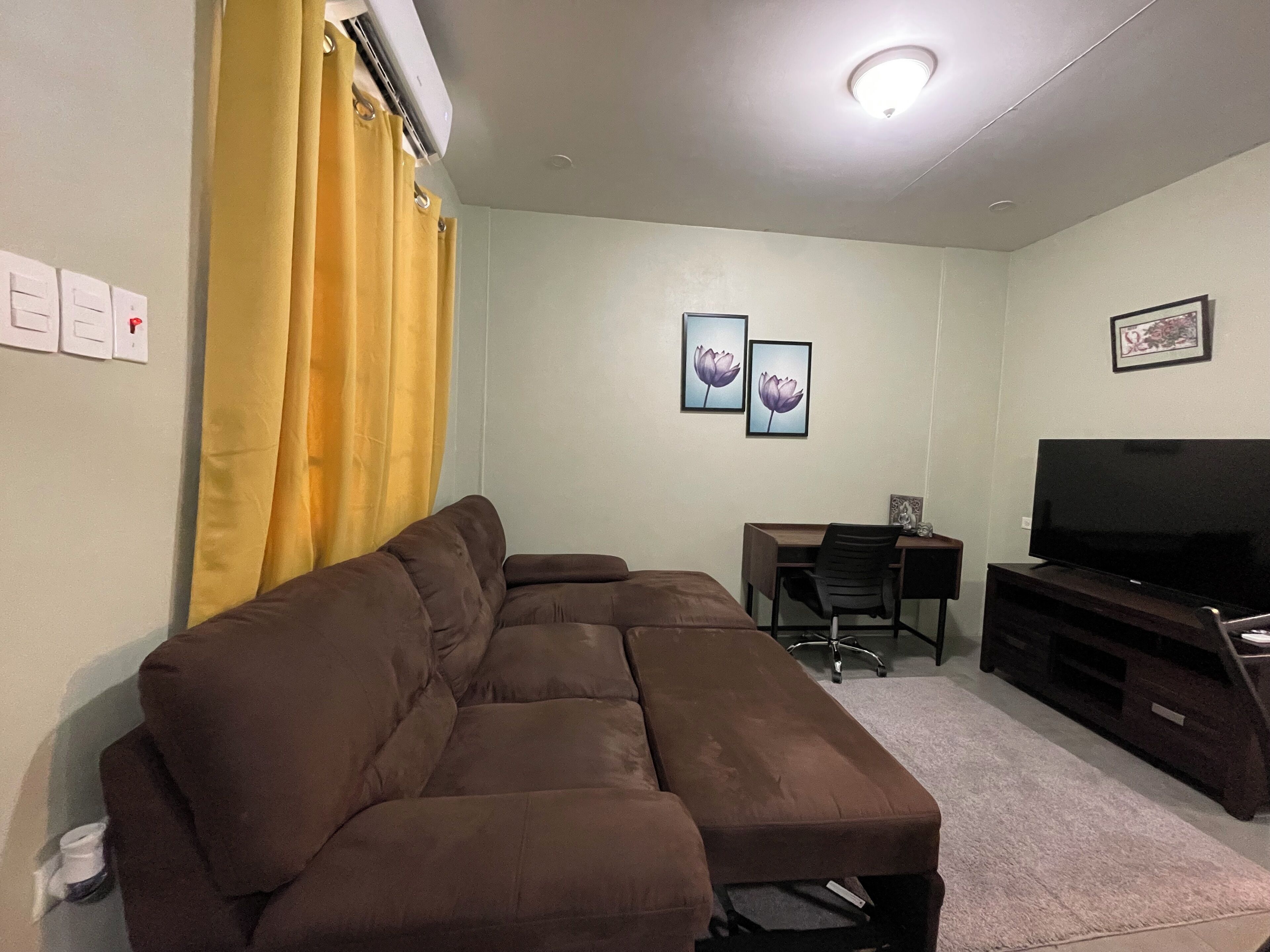 A living room with a brown sectional sofa, a flat-screen TV on a wooden stand, a desk with a chair, and two framed pictures on the wall.