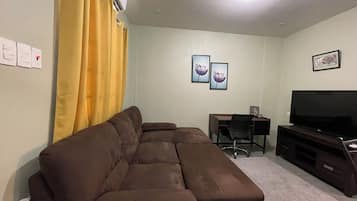 A living room with a brown sectional sofa, a flat-screen TV on a wooden stand, a desk with a chair, and two framed pictures on the wall.