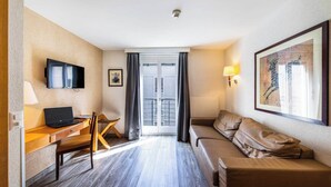 Une chambre d’hôtel avec un bureau en bois, un ordinateur portable, un canapé en cuir marron, un téléviseur fixé au mur et une œuvre d’art encadrée accrochée au mur.