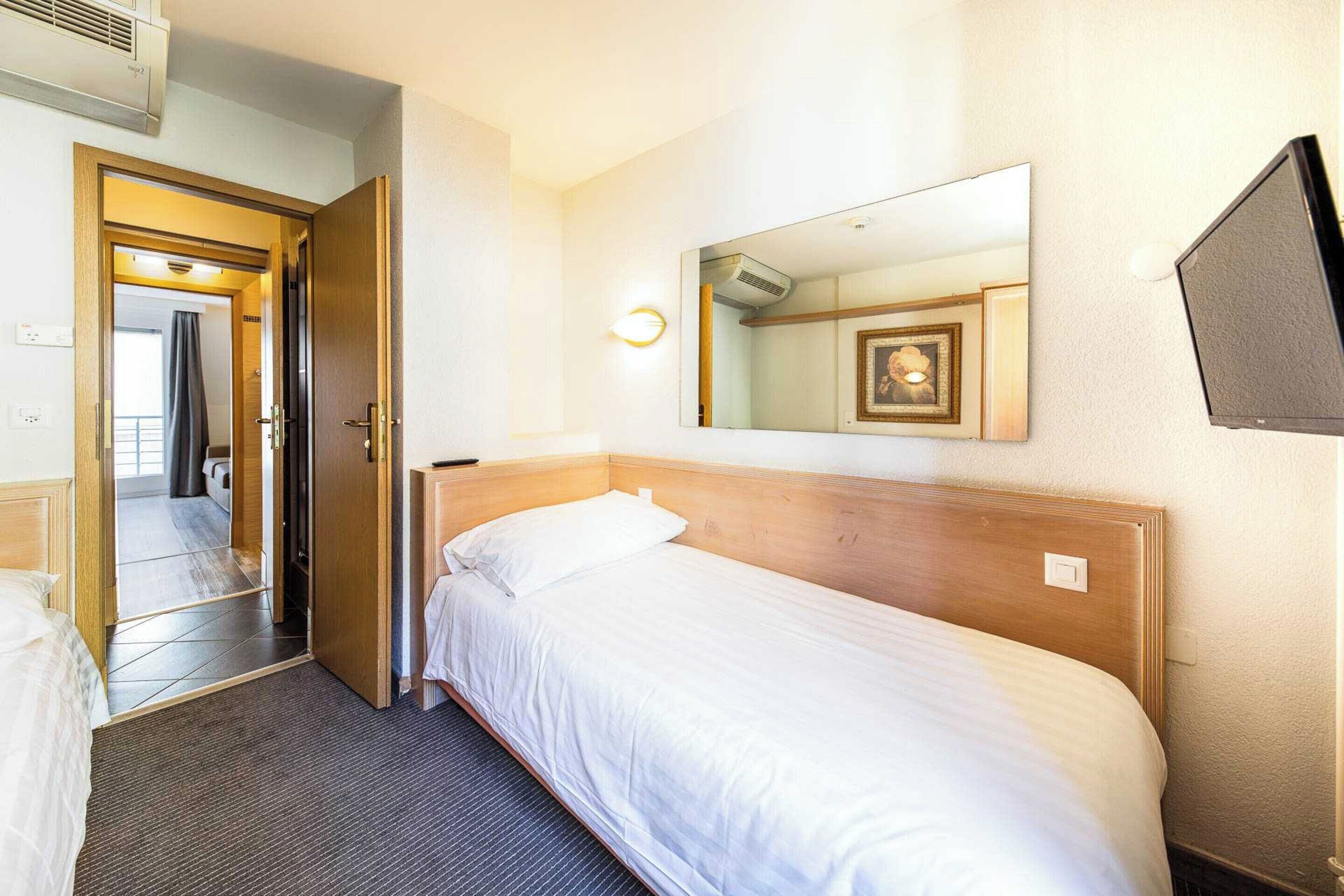 A hotel room with a single bed, a wooden headboard, a wall-mounted TV, and a mirror reflecting the room's interior.