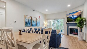 Una sala de estar moderna con chimenea, un comedor con mesa y sillas de madera, y un ventanal con vistas a árboles.