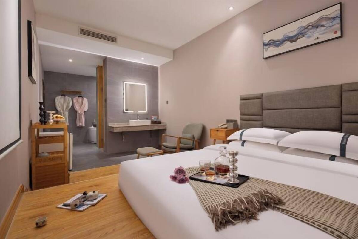 A hotel room with a large bed, a bedside table with a tray of drinks, a mirror, a chair, and a desk with a lamp.