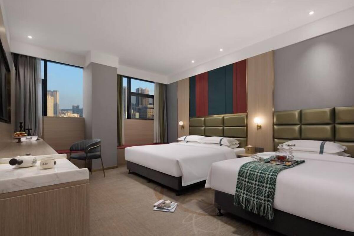 A hotel room with two beds, a desk, and a large window offering a city view.