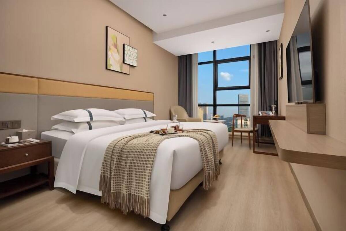A modern hotel room with a large bed, a desk, a chair, and a view of the city through large windows.