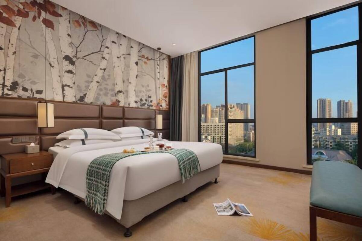 A hotel room with a large bed, a nightstand, a reading chair, and a city view through large windows.