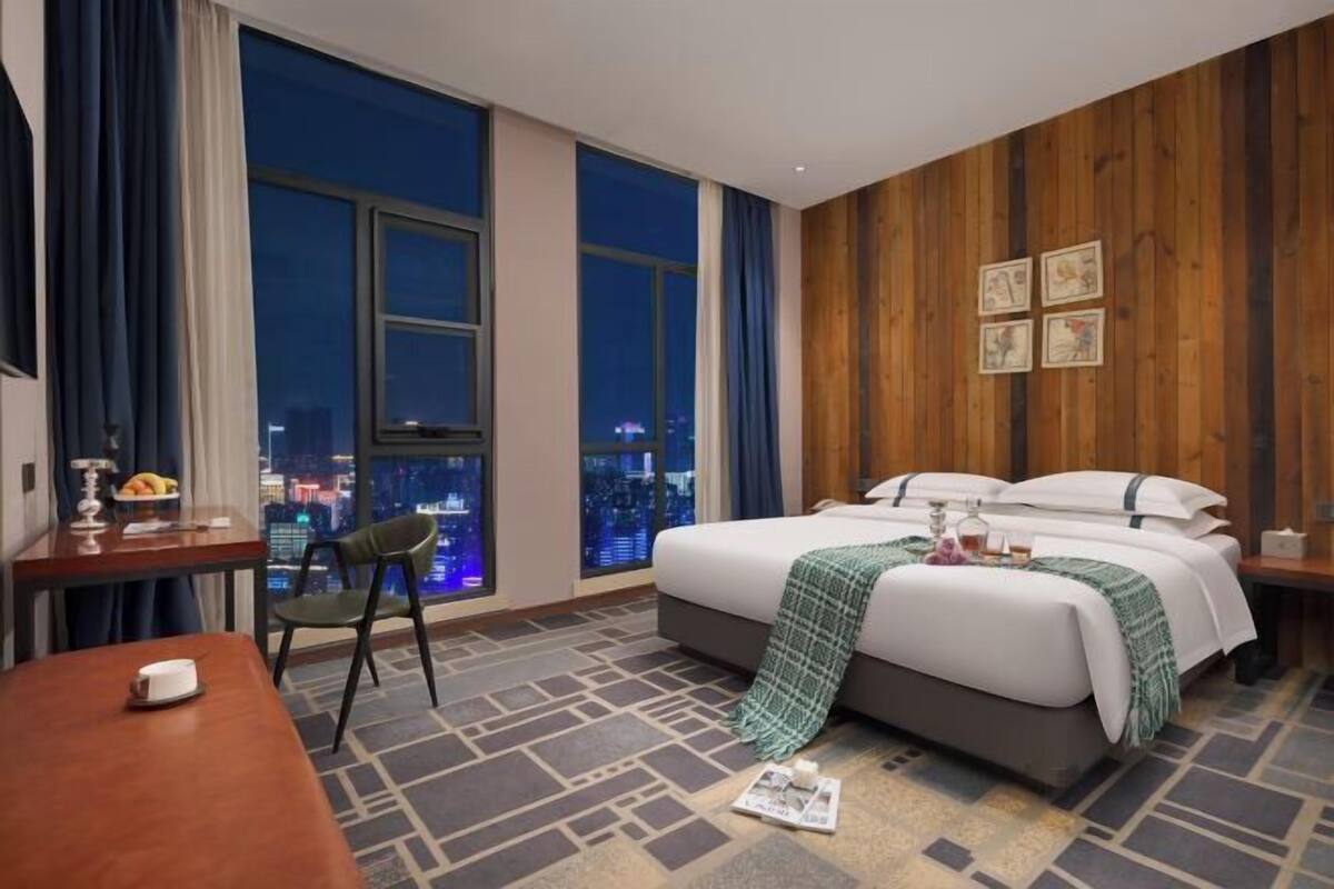 A hotel room with a large bed, a desk, a chair, and a view of the city at night.
