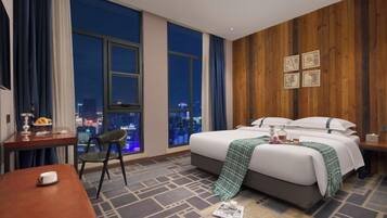 Habitación de hotel con una cama grande, un escritorio, una silla y vistas de la ciudad de noche.
