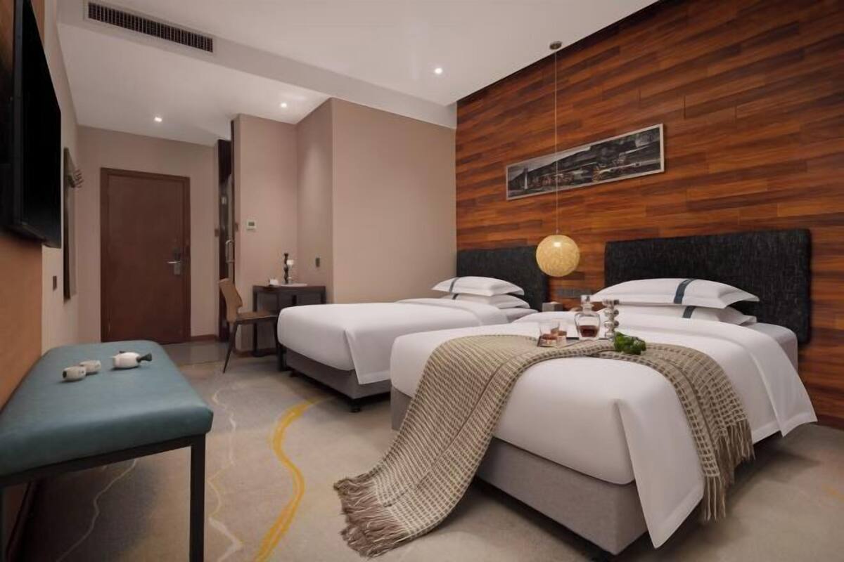 A hotel room with two beds, a bedside table, a bench, a wooden accent wall, and a mounted TV.