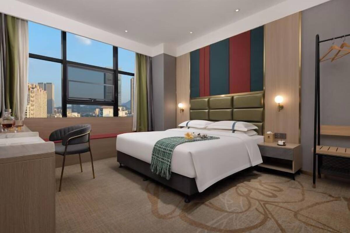 A hotel room with a bed, a desk, a chair, a nightstand, and a view of the cityscape.