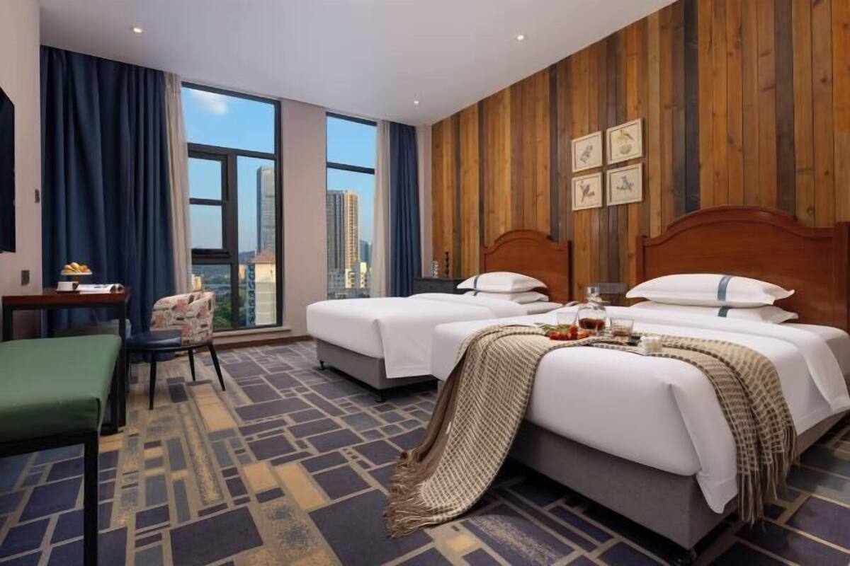 A hotel room with two beds, a desk, a chair, a large window with a city view, and wooden paneling on the walls.