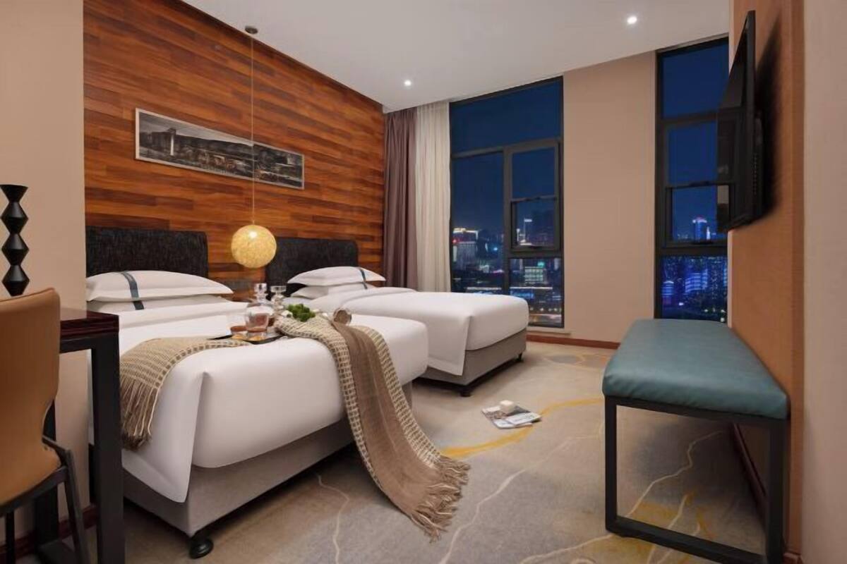 A hotel room with two beds, a desk, a chair, a TV, and a large window with a city view.