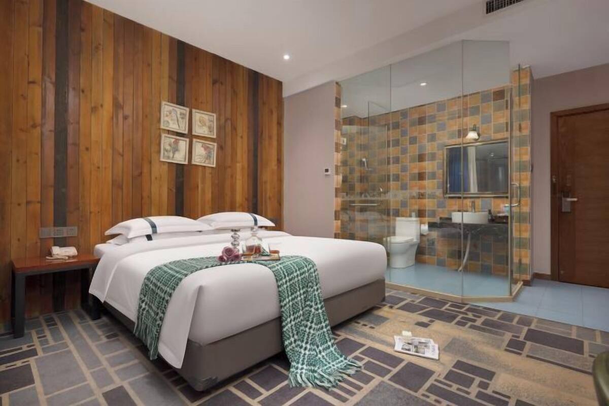 A modern hotel room with a large bed, a wooden accent wall, a glass shower enclosure, and a small bedside table.