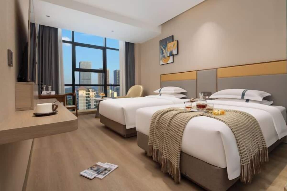A hotel room with two beds, a desk with a cup, a chair, and a large window with a city view.