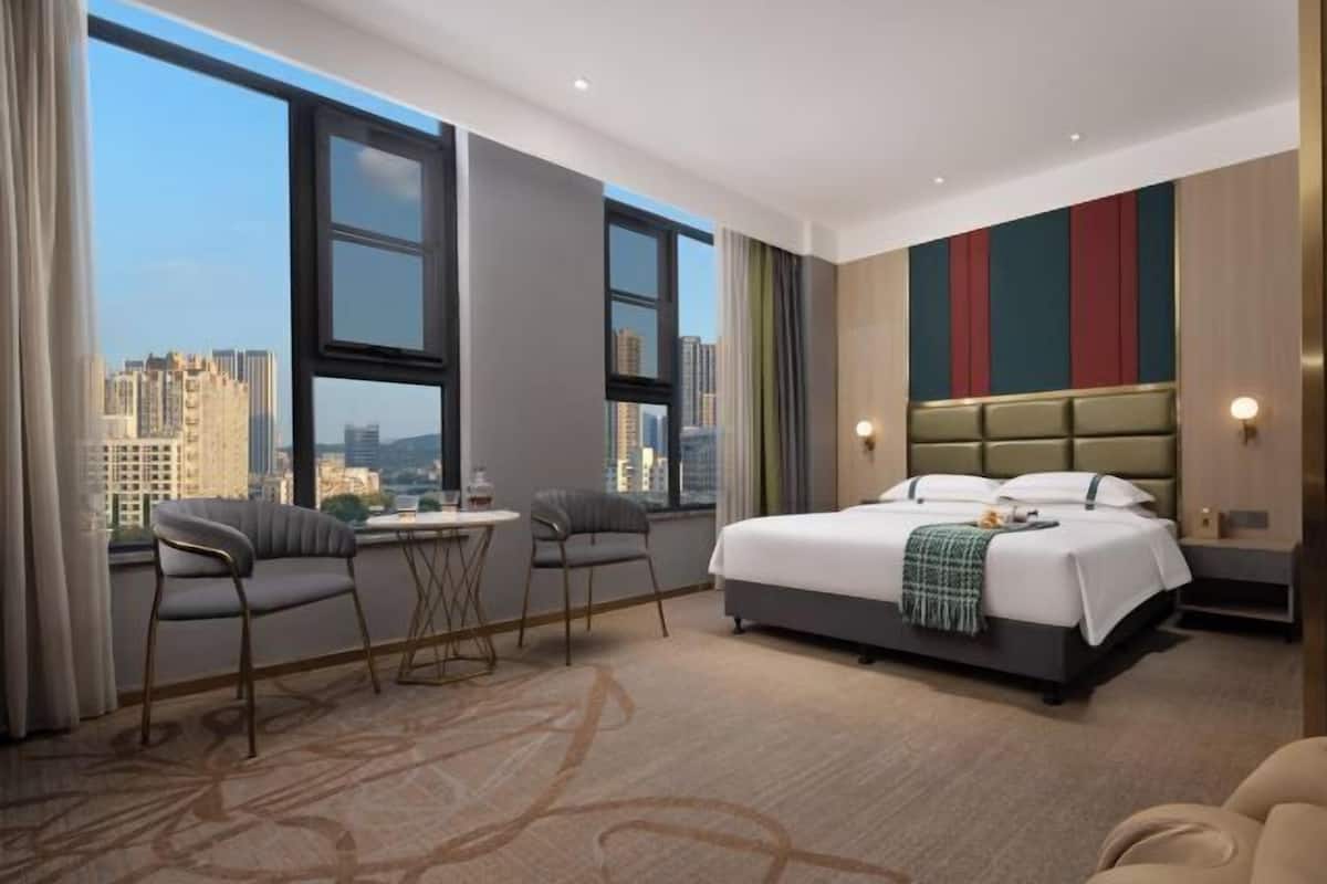 A hotel room with a large bed, two chairs, a small table, and a view of the cityscape.