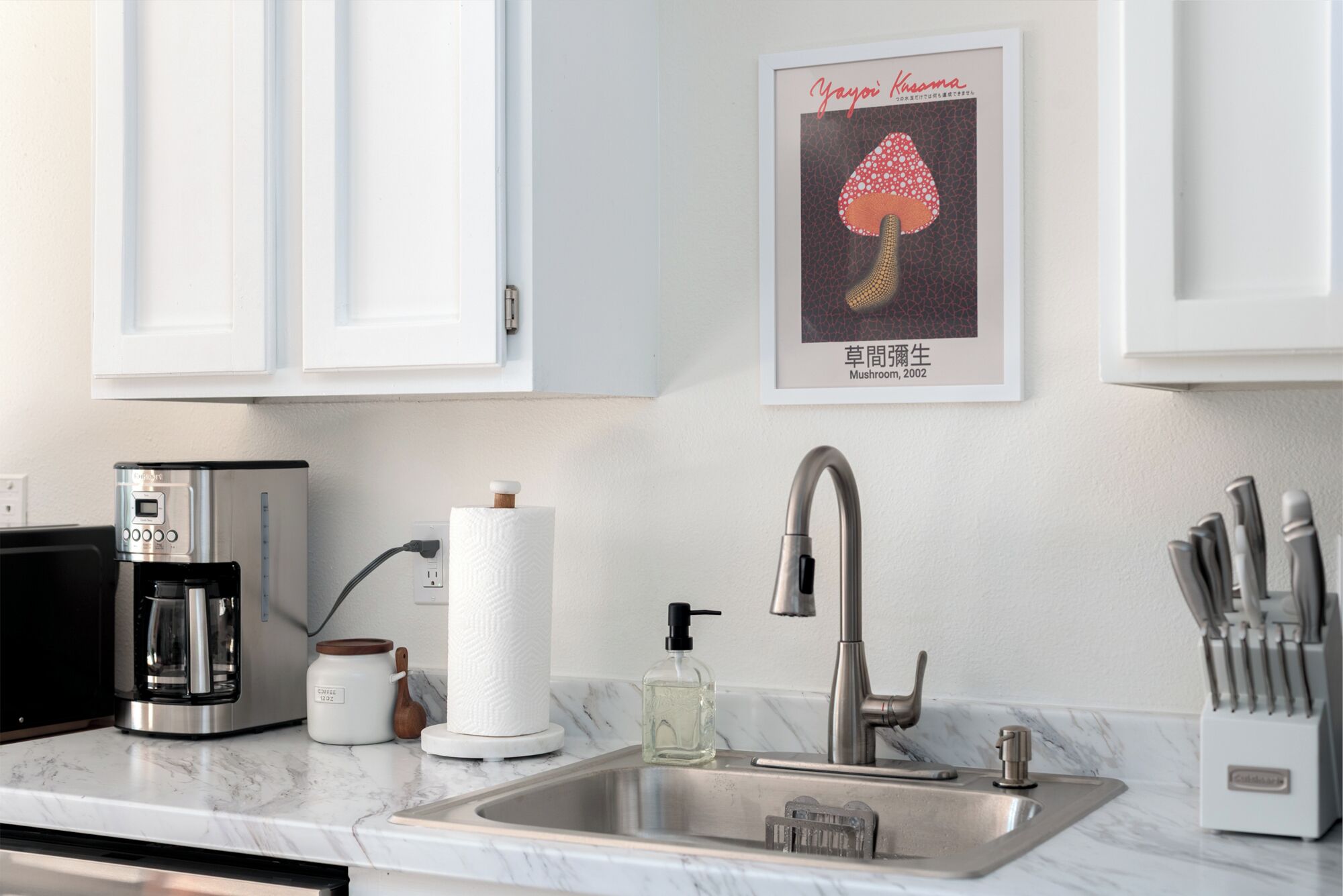 A kitchen with white cabinets, a stainless steel sink, a coffee maker, a paper towel holder, and a knife block.