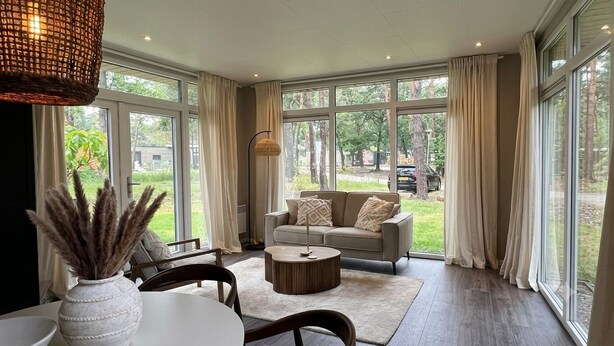 A spacious living room with a sofa, coffee table, and large windows offering a view of the outdoors. - Wellness Lodge Hottub on the Salland Ridge in Hellendoorn (Haarle)