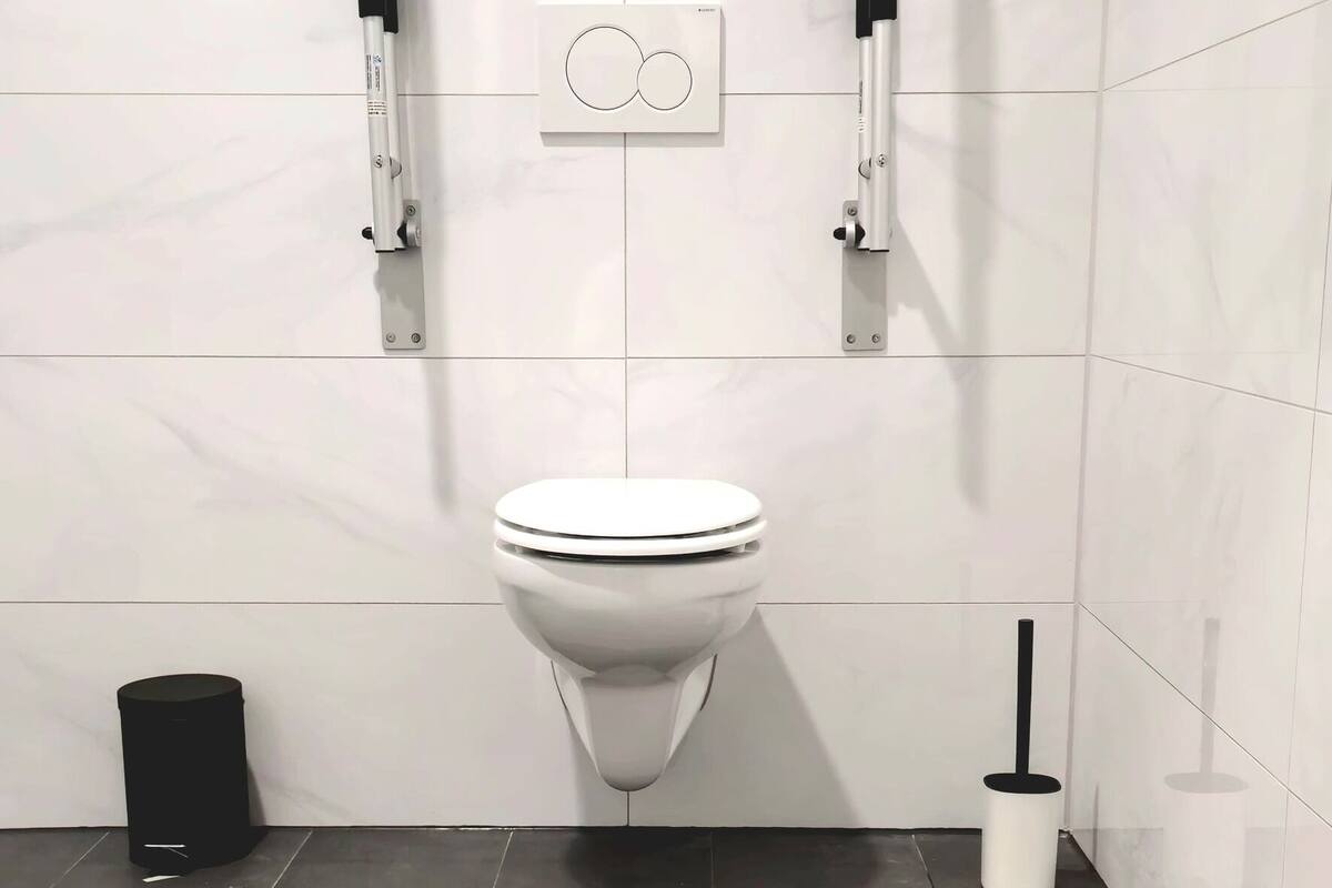 A modern bathroom with a white toilet, two black bidet handles, a black trash bin, and a white toilet brush.