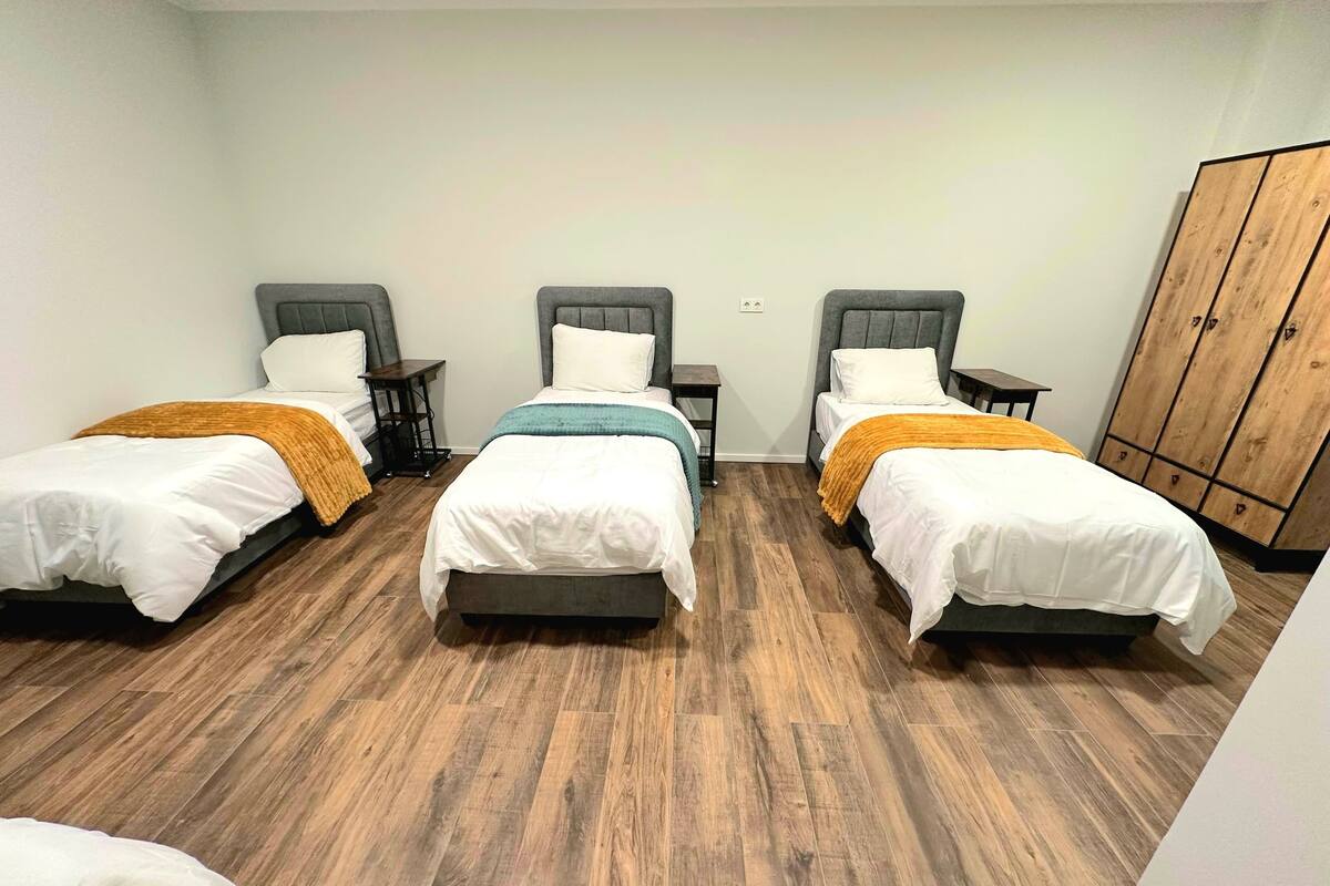 A room with three single beds, each with a wooden headboard, a small bedside table, and a nightstand. The room has a wooden floor and a wardrobe with a wooden finish.