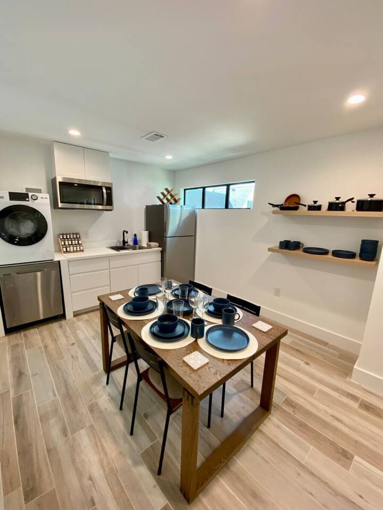 Cocina compacta con electrodomésticos modernos, comedor con mesa de madera para cuatro comensales y estanterías empotradas sobre la encimera.