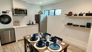Une cuisine compacte dotée d’appareils électroménagers modernes, un coin repas avec une table en bois pour quatre personnes, et des étagères intégrées au-dessus du plan de travail.