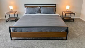 A double bed with a wooden frame and grey bedding, flanked by two bedside tables with lamps, in a room with a carpeted floor. - 4-BR Getaway Ideal for Families (Richmond)