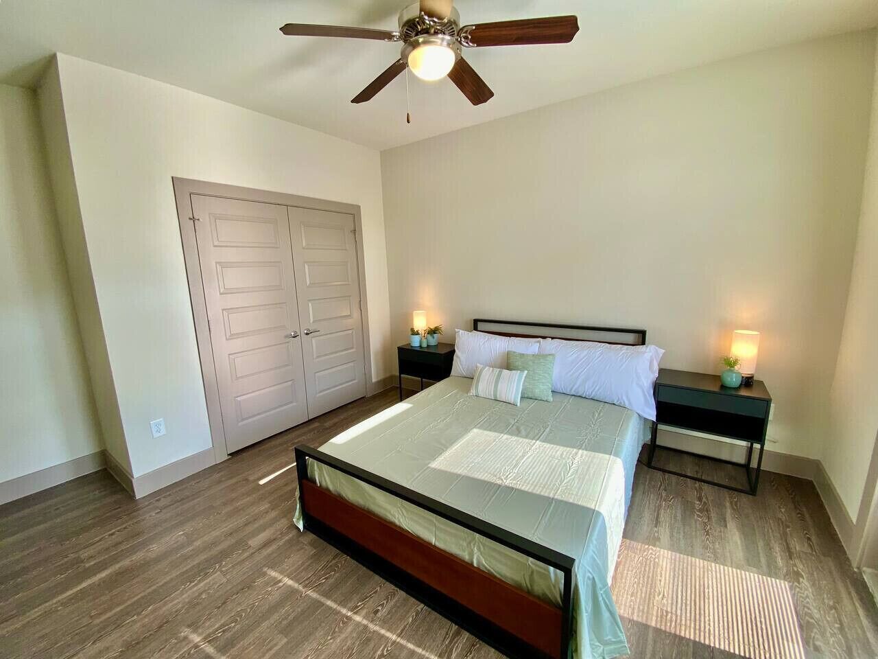 A bedroom with a bed, two bedside tables with lamps, a ceiling fan, and a door.
