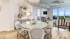 A dining area with a glass table surrounded by wicker chairs, a chandelier, and a view of the ocean through large windows.