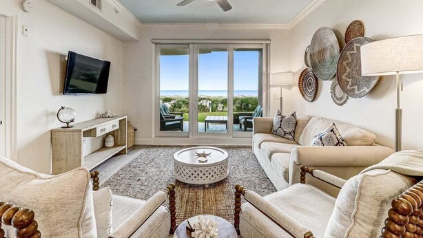 A spacious living room with a ceiling fan, a flat-screen TV, a sofa, a coffee table, and a view through large glass doors to the outdoors.
