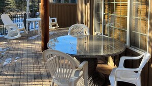 Een overdekt terras buiten met een ronde glazen tafel en witte kunststof stoelen, omgeven door houten terrastegels en een houten omheining.