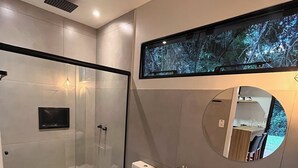 A modern bathroom with a glass shower enclosure, a toilet, a sink with a black countertop, and a mirror with a circular frame. - Rastro Cabins (Restinga Sêca)