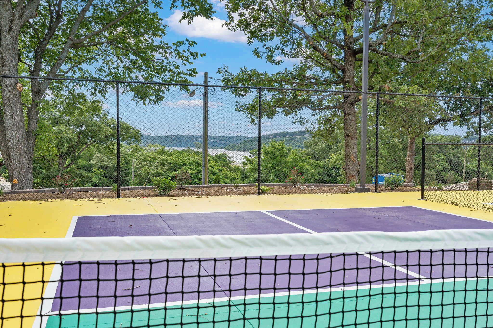 Una cancha de tenis con una red al frente, rodeada por una cerca de malla y árboles.