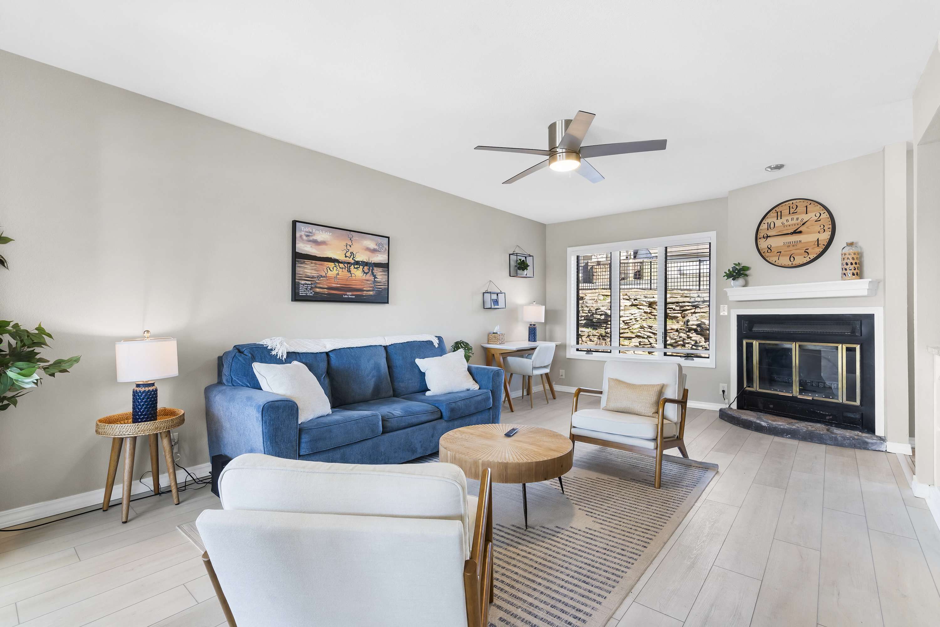 A living room with a blue sofa, white armchairs, a wooden coffee table, a fireplace, a clock, and a ceiling fan.