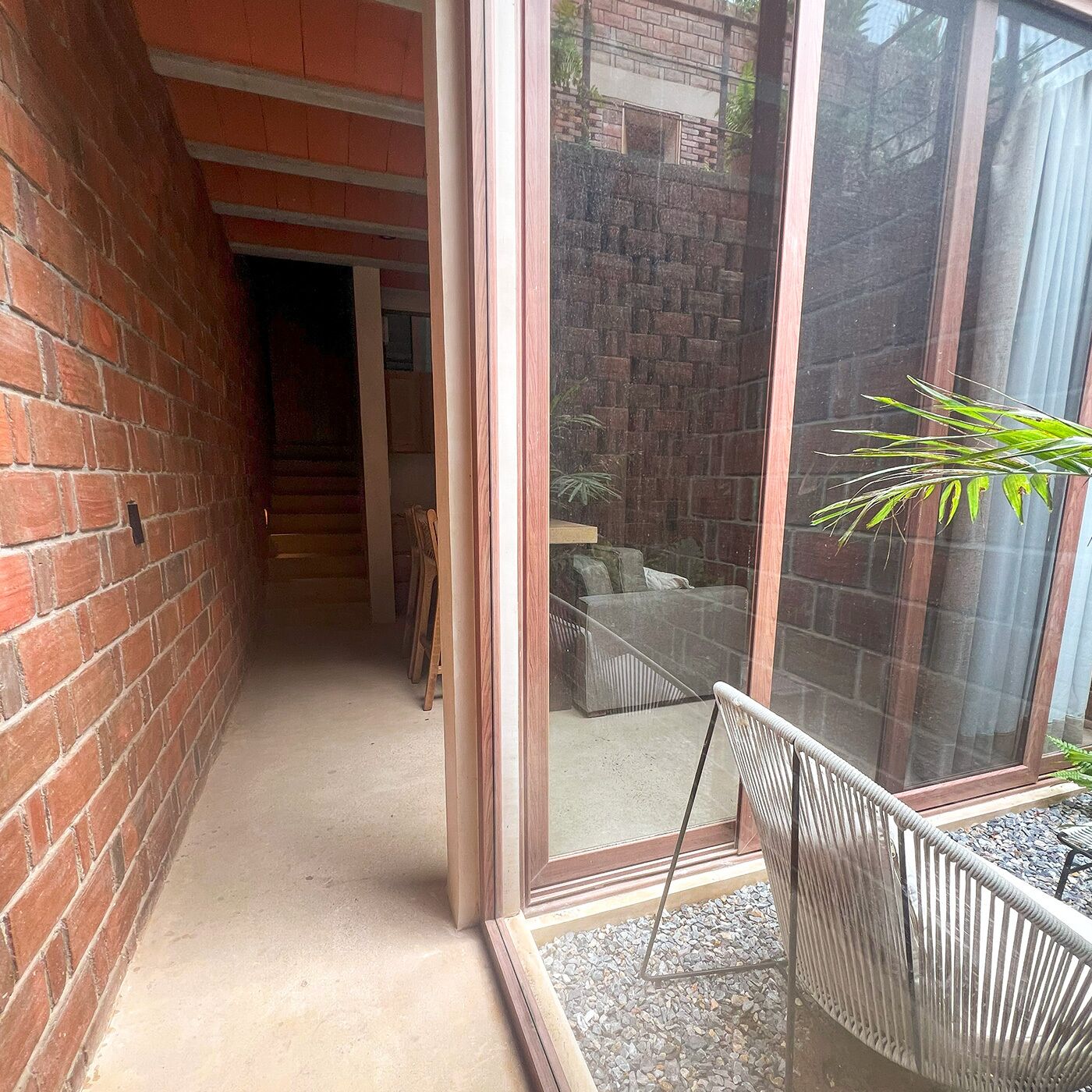 Un estrecho pasillo con pared de ladrillo, puerta de vidrio que da a un patio con mobiliario y una planta.
