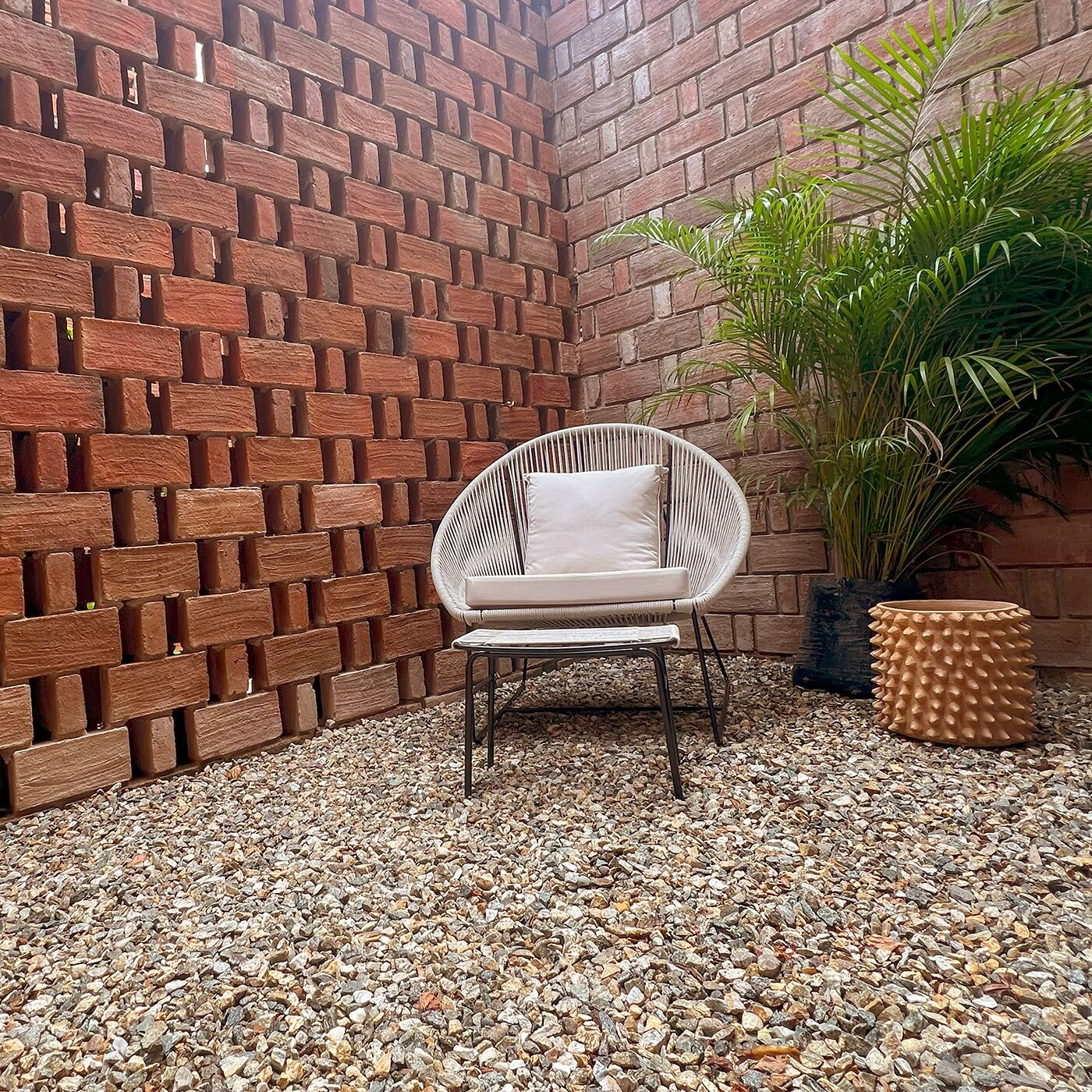 Una silla de mimbre con cojín, una planta en maceta y una mesa redonda con textura en una esquina, junto a una pared de ladrillo y suelo de grava.