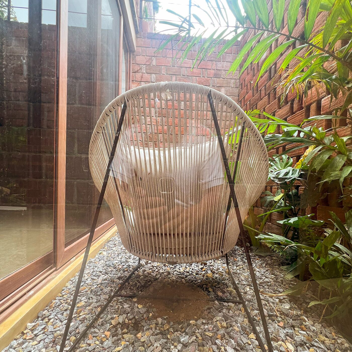 A wicker chair with a metal frame, positioned on a gravel surface, adjacent to a brick wall and a glass door.
