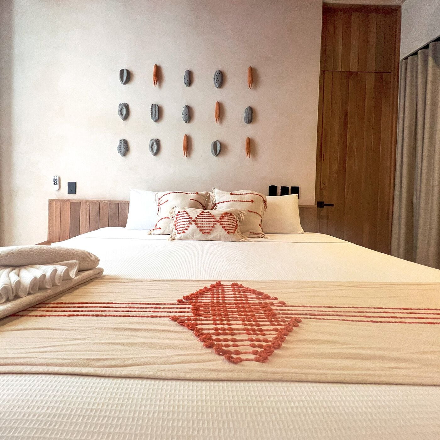 A neatly made bed with a white and red patterned bedspread, a wooden headboard, and a wall decorated with various textured wall hangings.