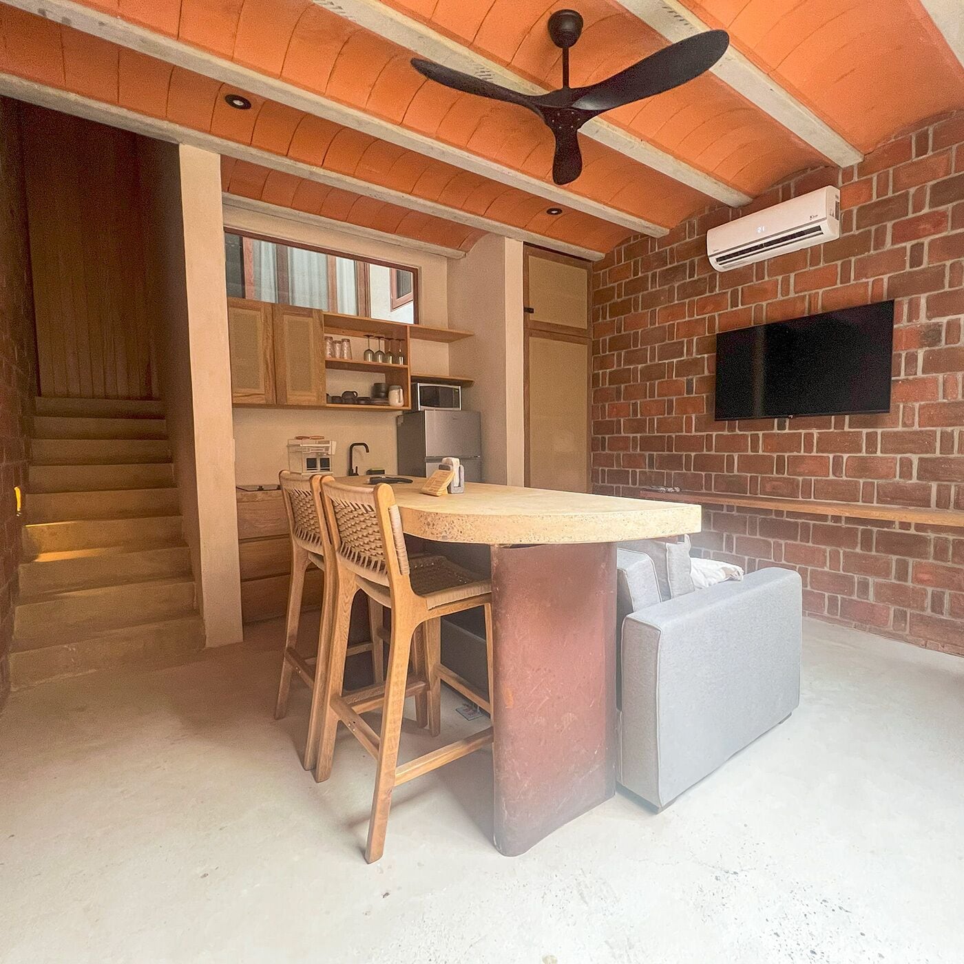 A kitchen with a dining area, wooden chairs, a round table, a ceiling fan, a TV mounted on a brick wall, and a staircase.