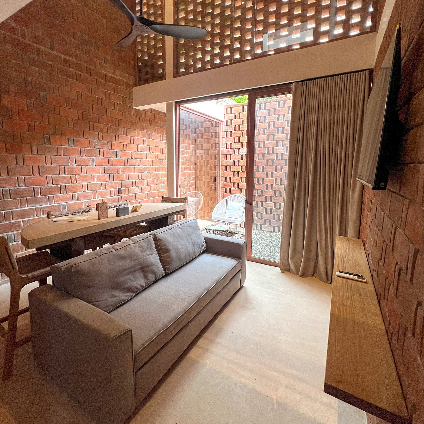 A modern living room with a brick wall, a wooden dining table, a sofa, and a TV mounted on the wall.