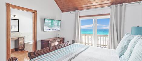 A bedroom with a wooden ceiling, a bed with a striped bedspread, a dresser, a television on the wall, and a view of the ocean through a balcony door.