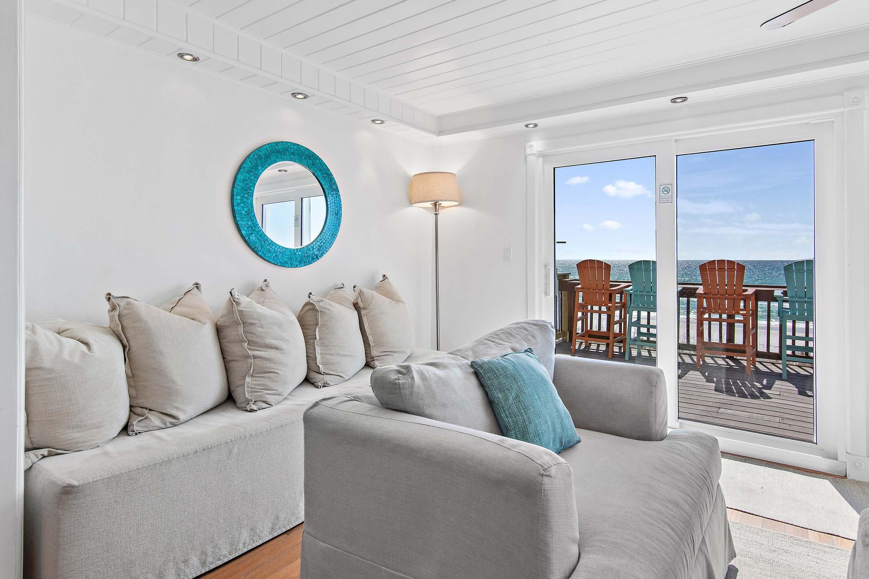 Uma sala de estar moderna com um sofá cinza, um espelho redondo e vista para o deck e oceano.
