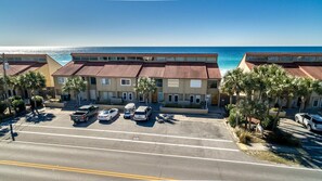 A row of two-story buildings with red-tiled roofs, a parking lot with several cars, and a view of the ocean. - Green Reef #8- Beachy Keen (Destin)