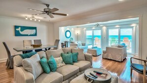 Un salon spacieux avec un canapé beige, des coussins turquoise, une table basse avec des télécommandes, et un coin repas comprenant une table et des chaises.