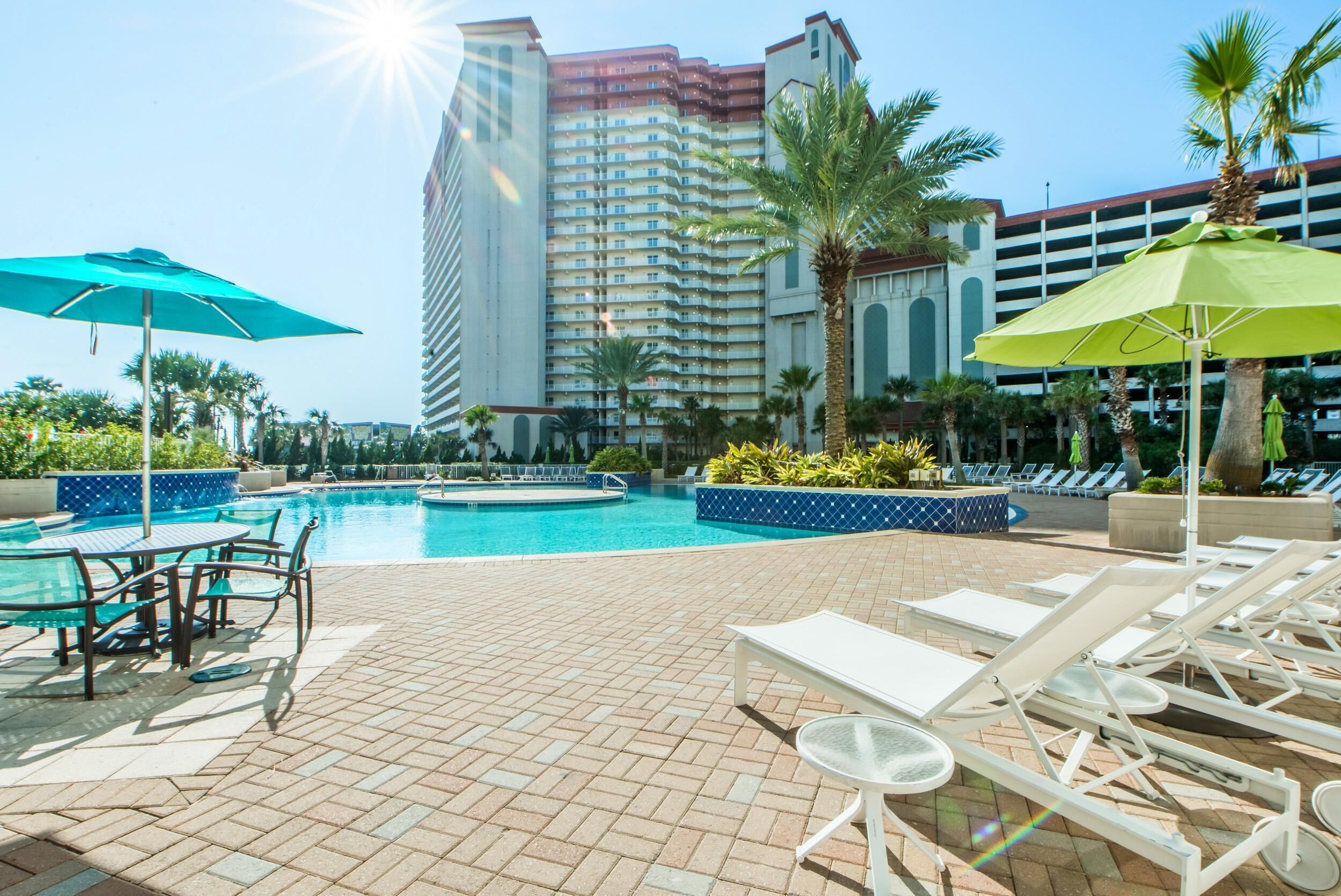 A hotel pool area with lounge chairs, umbrellas, and a large building in the background.