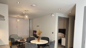 Ein modernes Wohnzimmer mit Essbereich, ausgestattet mit einem runden Tisch, grau gepolsterten Stühlen, einem Fernseher und einer dekorativen Vase.